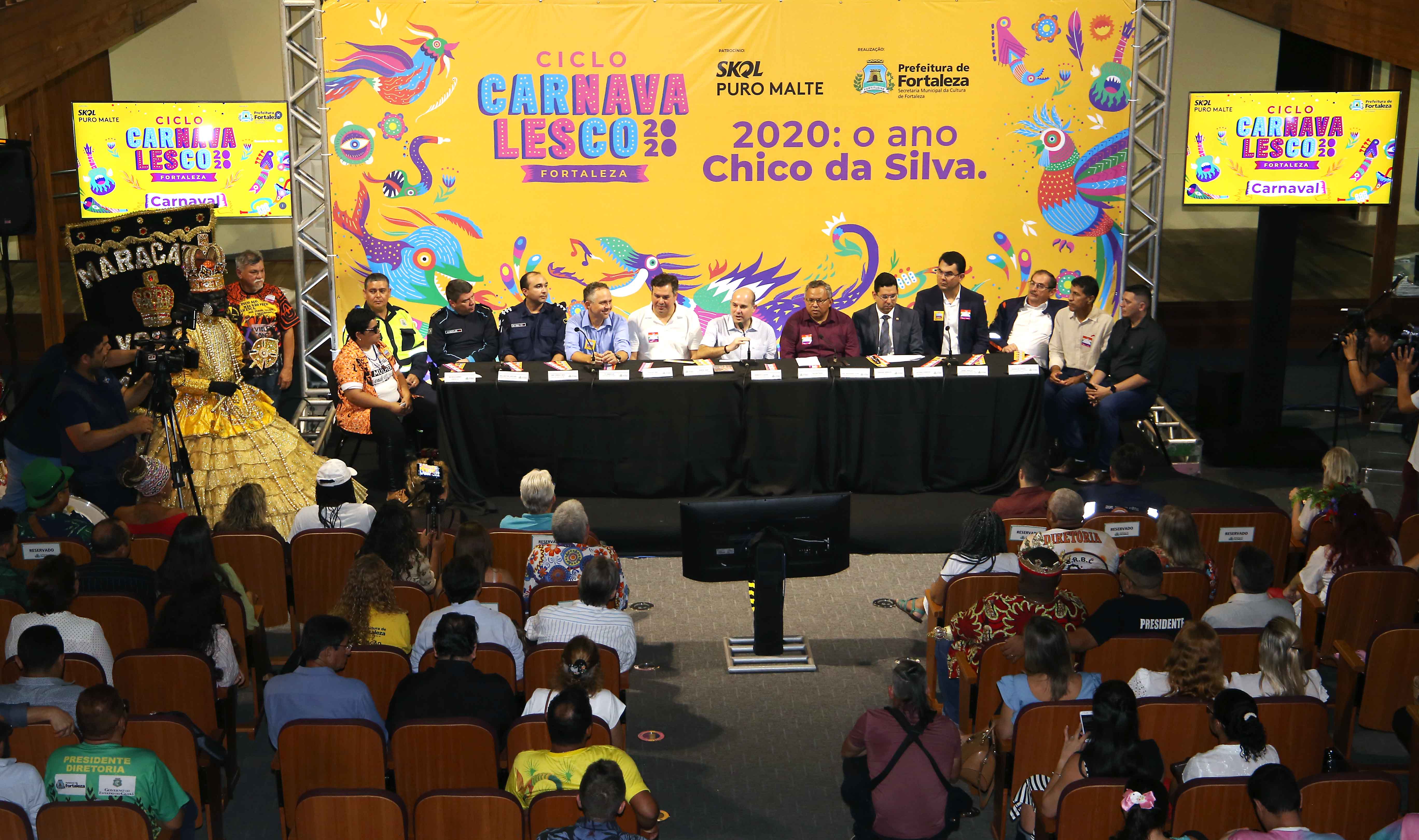 prefeito e pessoas sentadas à mesa com público sentado em frente assistindo